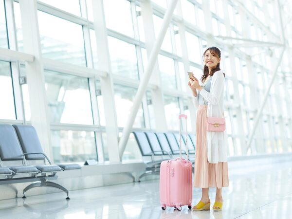 空港でピンク色のスーツケースとスマホ片手のロングスカートの女性